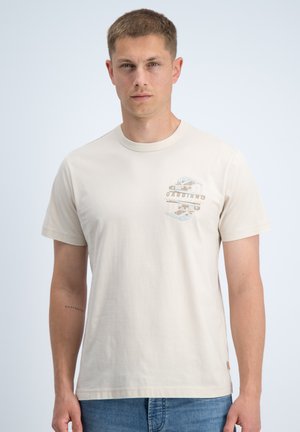 Jonge man met kort haar die een licht beige T-shirt met een klein borstmotief en blauwe spijkerbroek draagt, staand tegen een effen achtergrond.