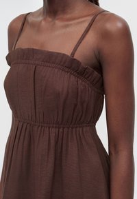 Femme portant une robe marron sans manches avec de fines bretelles et des détails froncés au niveau de la poitrine et de la taille.