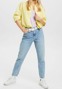 Žlutá košile na knoflíky, levandulový top, světle modré džíny, bílý pásek a bílé sneakers. Modelka stojí sebevědomě, s rukama na bocích.