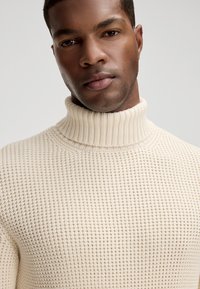 Kremowy sweter z golfem w teksturowanym wzorze dzianiny, z prążkowanym kołnierzem i gładką sylwetką.