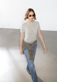 Top corto grigio chiaro a maniche corte, abbinato a jeans blu a palazzo. Un maglione a righe marroni e neri annodato in vita. Occhiali da sole marroni indossati.