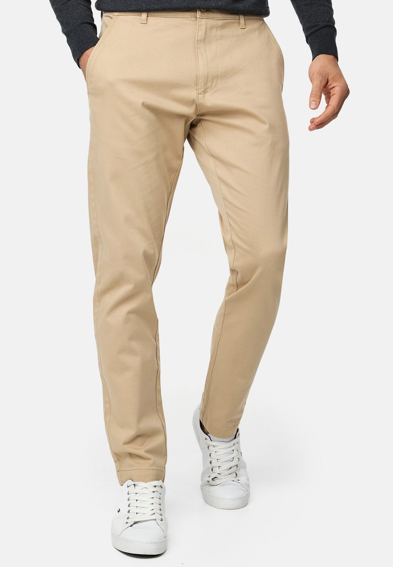 Uomo che indossa chinos beige slim-fit con le mani nelle tasche e sneaker bianche, in piedi davanti a uno sfondo semplice.