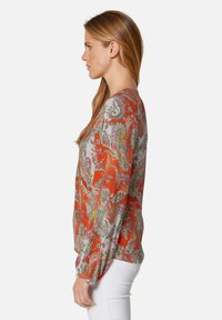 Langärmlige Bluse in Orange mit einem Paisleymuster in Grün und Pink. Das Material scheint leicht und hat eine glatte Textur sowie elastische Bündchen.