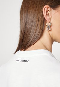 Pendientes con un diseño en oro y plata, con una forma de bucle entrelazado. El modelo lleva una camisa blanca con "KARL LAGERFELD" impreso en la parte posterior.