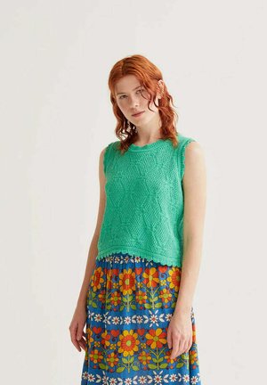 Joven con cabello rojo que lleva una blusa verde sin mangas de punto y una falda azul con flores naranjas y amarillas.