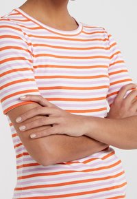 Pruhované tričko s krátkým rukávem, které má horizontální pruhy v oranžové, růžové a bílé barvě. Kulatý výstřih a měkká bavlněná látka.