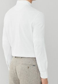 White long-sleeve shirt with a small collar, paired with light brown herringbone trousers featuring a button and a small brand label.