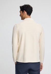 Koszula polo z długim rękawem w kolorze kremowym z klasycznym kołnierzykiem. Gładki materiał, prosty krój i ściągacze na rękawach. Model stoi z rękami w kieszeniach.