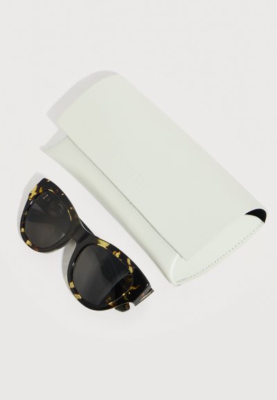 Γυαλιά ηλίου με σκελετό σε σχήμα γατιού σε σχέδιο χελώνας και σκουρόχρωμους φακούς, δίπλα σε μια λευκή ορθογώνια θήκη γυαλιών Burberry πάνω σε λευκή επιφάνεια.