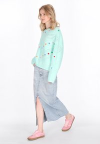 Maglione verde menta con motivi di cuori multicolori, abbinato a una gonna di denim chiaro con uno spacco laterale e scarpe rosa con suola marrone.