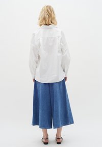 White, long-sleeve shirt with a relaxed fit and back yoke detail, paired with a blue denim culotte skirt. Smooth texture, modern design.