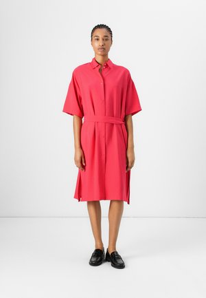 Button-up shirt dress in bright pink fabric. Features short sleeves, a belted waist, side slits, and a relaxed fit. Paired with black loafers.