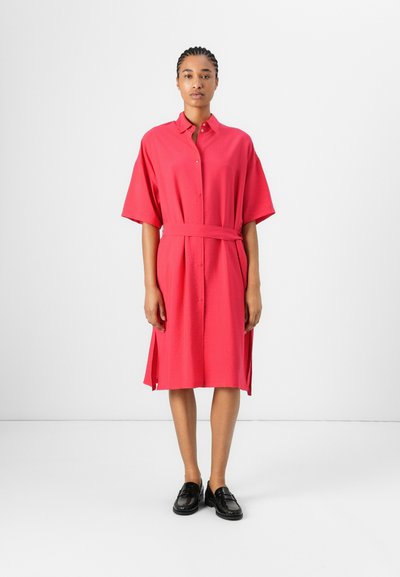 Robe chemise à boutons en tissu rose vif. Caractéristiques des manches courtes, une taille ceinturée, des fentes latérales et une coupe décontractée. Associée à des mocassins noirs.