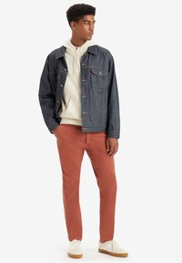 Veste en denim avec des coutures claires, superposée sur un sweat-shirt blanc, accompagnée de pantalons couleur rouille et de sneakers blanches avec des accents beiges.
