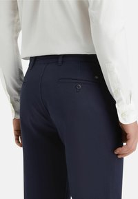 Navy blue trousers made of smooth fabric featuring a back pocket with a button, paired with a white button-up shirt.