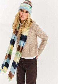 Beżowy sweter z grubym splotem, ciemnobrązowe spodnie oraz wielokolorowy szalik w kratę. Niebieska i brązowa dzianinowa czapka w paski z pomponem.