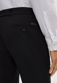 Zwarte broek van gladde stof met een enkele achterzak met een knoop, vergezeld van een rechthoekig logopatch.