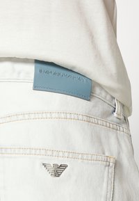 Světlomodrý kožený štítek s nápisem "Emporio Armani" na bílých denimových džínách, s kovovým logem a kontrastním žlutým prošitím.