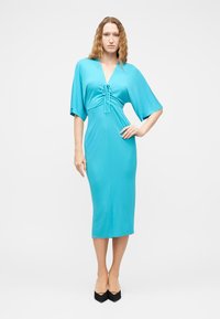 Γυναίκα με μακριά κυματιστά μαλλιά, φορώντας ένα τιρκουάζ μεσαίο φόρεμα με φαρδιές μανίκες και ραμμένη μπροστινή πλευρά, στέκεται με το ένα χέρι στη μέση και μαύρες γόβες.