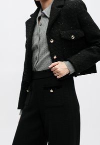 Femme portant un blazer noir texturé avec des boutons dorés sur un cardigan gris en maille torsadée et un pantalon noir taille haute assorti avec des détails de boutons dorés.