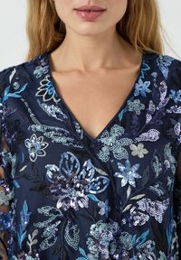 Blusa floral azul marino con delicado bordado de cuentas y lentejuelas en tonos azules, con escote en V y mangas transparentes.