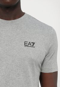 Mann iført lysegrå kortermet T-skjorte med svart EA7 Emporio Armani-logo på venstre bryst.