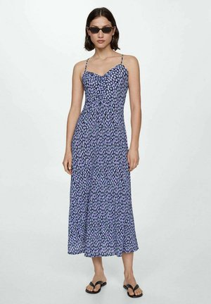 Femeie îmbrăcată într-o rochie florală violet, fără mâneci, până la gleznă, cu sandale negre tip flip-flop, cercei rotunzi și ochelari de soare rectangulari negri, stând în fața unui fundal simplu.