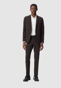 Costume marron foncé avec une coupe ajustée, fermeture à un bouton, chemise blanche et chaussures noires. Tissu lisse et plis marqués visibles.