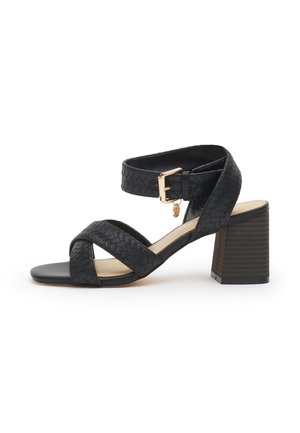 Sandalo in pelle nera intrecciata con tacco largo a blocco, cinturino alla caviglia e fibbia color oro con piccolo ciondolo.