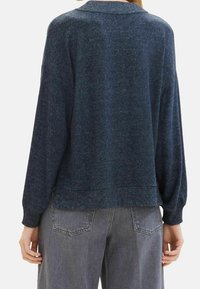 Väljä tummansininen neulepusero, jossa on teksturoitu neulos ja resorikaulus, pitkät hihat ja suora helma, yhdistetty harmaisiin housuihin.