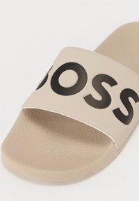 Beige Rutschsandale mit einem breiten Riemen und einem markanten schwarzen Logo, strukturiertem Fußbett und glatter Oberfläche. Ideal für Freizeitkleidung.