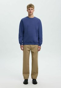 Mėlynas medvilnės džemperis su apvalia kaklo iškirpte ir megztomis rankovėmis, derinamas su smėlio spalvos tiesiomis kelnėmis ir juodais batais.