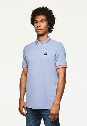 Homme aux cheveux bouclés portant un polo bleu clair avec des bordures orange sur le col et les manches, associé à un jean bleu, se tenant devant un fond blanc.