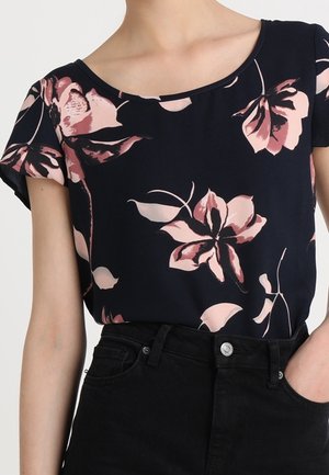 Zwart topje met korte mouwen en grote roze en beige bloemenprint, gedragen in een hoge zwarte jeans met een zilveren knoop.