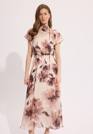 Kobieta ubrana w różowo-brązową, kwiatową sukienkę midi z krótkimi rękawami, z dopasowanym różowym chokerem i pierścionkiem, stojąca na jednolitym tle.