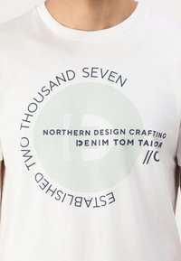Hvit bomull t-skjorte med rund hals, med en stor sirkulær grafikk i mintgrønn og marineblå, som viser tekst og en logo i midten.