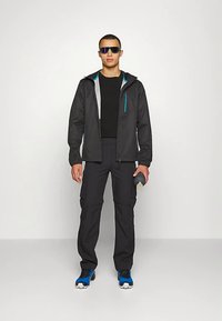 Svart vanntett jakke med blå detaljer, kombinert med svarte bukser og blå sko. Modellen holder en vannflaske og har på seg solbriller.