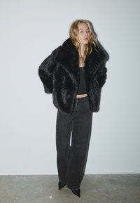 Sort jakke i imiteret pels med brede slag, båret over en tætsiddende sort top. Kombineret med strukturerede sorte bukser med diskrete pyntedetaljer og spidse hæle.