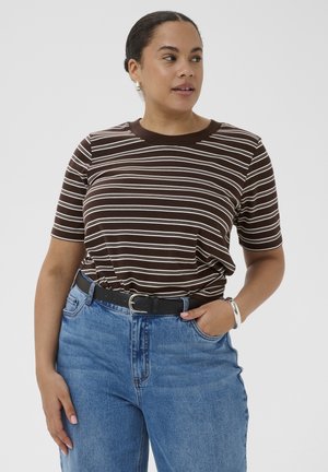 Kobieta w brązowo-białej paskowanej koszulce z krótkim rękawem włożonej do wysokich niebieskich dżinsów z czarnym paskiem, stojąca z jedną ręką w kieszeni.