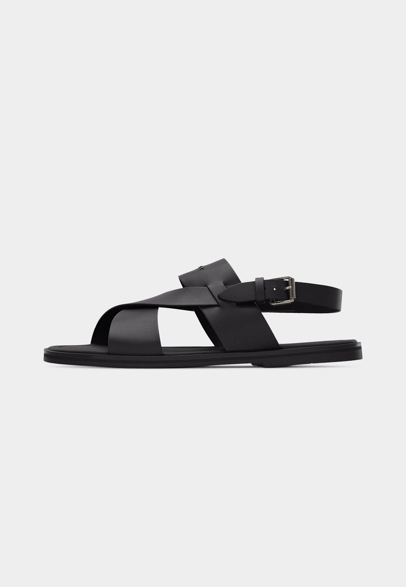 Μαύρο δερμάτινο σανδάλι με φαρδιές λουρίδες που διασταυρώνονται και ρυθμιζόμενο λουράκι αστραγάλου με πόρπη, απεικονισμένο σε απλό φωτεινό φόντο.