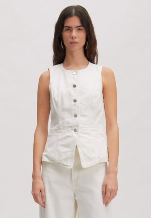 Vrouw met lang donker haar, gekleed in een mouwloos wit vest met knopen en bijpassende witte broek, staand tegen een effen lichte achtergrond.