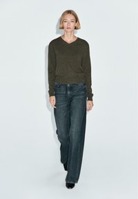 Ciemnozielony sweter w serek połączony z niebieskimi dżinsami o wysokim stanie i szerokich nogawkach. Modelka nosi czarne szpilki, stojąc na jasnym tle.