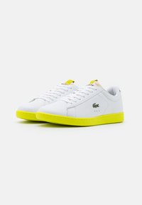 Vita lädersneakers med en texturerad yta, ljusgul sula och snörning. Har en grön logotyp på sidan och en gul accent på hälkappan.