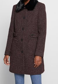 Cappotto bordeaux con texture e colletto in pelliccia sintetica nera, dotato di chiusura a bottoni, due tasche e maniche lunghe.