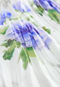 Ziedu plisētā kleita, kas ražota no auduma ar lieliem purpura ziediem un zaļām lapām uz balta fona, ar mīkstu, plūstošu tekstūru.