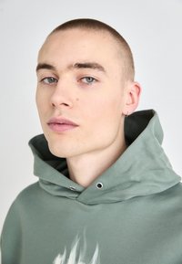 Sudadera verde con capucha ajustable, que presenta ojales de metal plateado y un diseño gráfico sutil en el pecho, con una textura de tela suave.