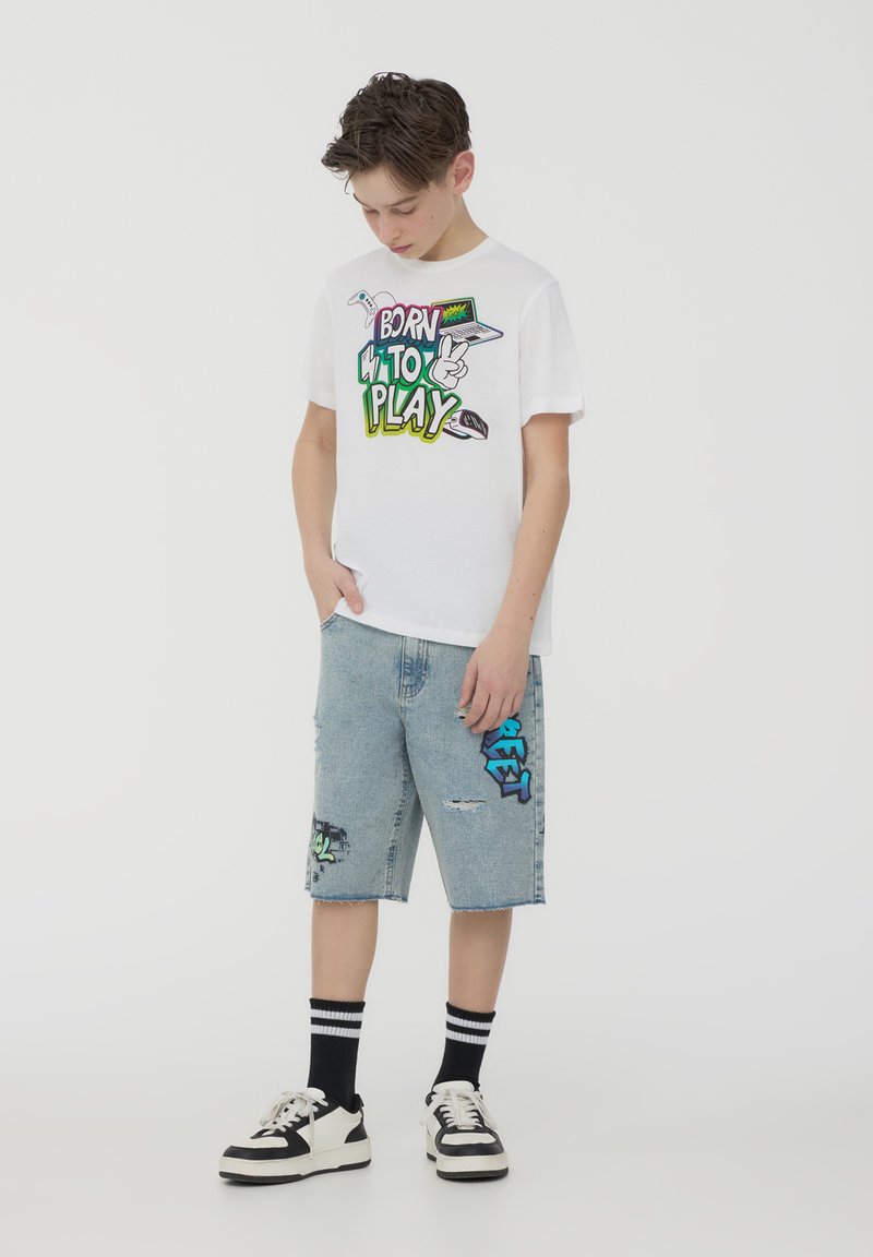 T-shirt bianco a maniche corte con grafica colorata "BORN TO PLAY". Pantaloni corti in denim azzurro chiaro con accenti in stile graffiti. Sneakers bianco e nere.