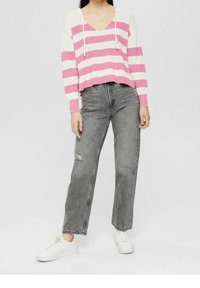 Mujer vestida con un suéter de punto a rayas rosa y blanco, jeans grises de tiro alto y rasgados, y zapatillas blancas, de pie contra un fondo liso.