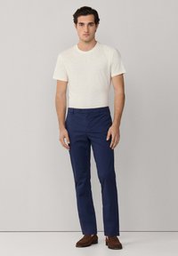 Mand står mod en ensfarvet baggrund iført en hvid kortærmet skjorte, marineblå bukser og brune loafers.