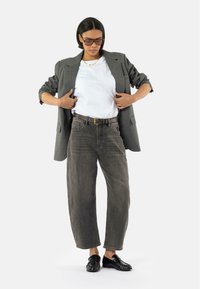 Blazer grigio, t-shirt bianca e jeans grigi a vita alta abbinati a mocassini neri. Il blazer presenta spalline imbottite e due tasche frontali.